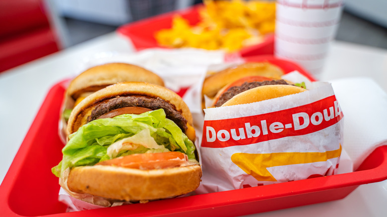Ένα in-n-out διπλά διπλά μπιφτέκια κάθονται σε ένα κόκκινο δίσκο εστιατορίων μπροστά από τηγανητές πατάτες και ένα ποτό στο κλασικό φλιτζάνι φοίνικα του