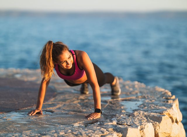 Καλή νεαρή γυναίκα που κάνει ασκήσεις push-up κατά τη σκληρή εκπαίδευση κοντά στην παραλία της θάλασσας