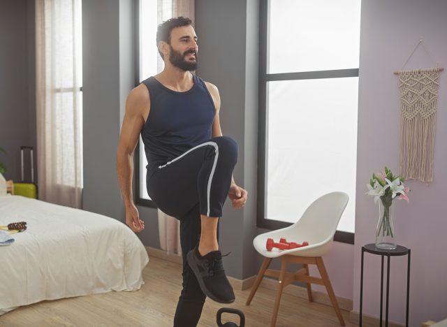 Ένας γενειοφόρος άνδρας σε αθλητική ενδυμασία που ασκεί σε ένα καλά φωτισμένο, μοντέρνο υπνοδωμάτιο, ανυψώνοντας το γόνατό του για μια ρουτίνα προπόνησης.