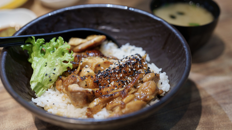 Ένα μπολ με κοτόπουλο teriyaki γαρνιρισμένο με σουσάμι πάνω από ρύζι, με σούπα miso στο παρασκήνιο